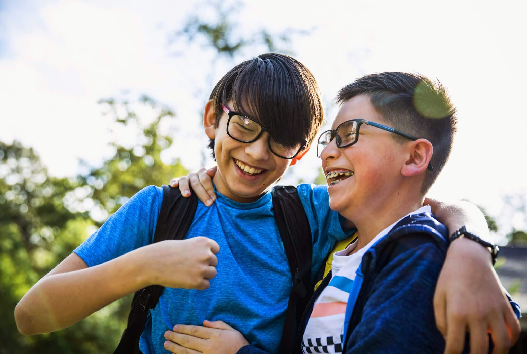 Two brothers wearing backpacks and glasses hug and laugh together